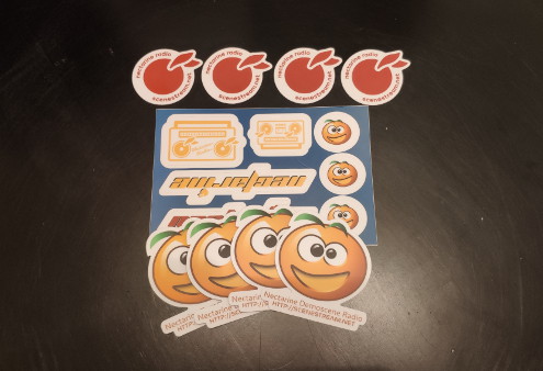 Corner of a black table with one sticker sheet, four large Necta stickers, and four fruit stickers with text around the edges arranged on top