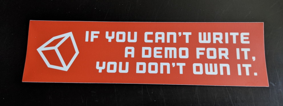 A large rectangular bumper sticker with the text 'If you can’t write a demo for it, you don’t own it' and have a cube on it