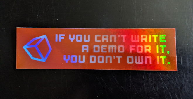 A small rectangular holographic sticker with the text 'If you can’t write a demo for it, you don’t own it' and have a cube on it
