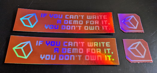 Two small rectangular holographic stickers that say 'If you can’t write a demo for it, you don’t own it' and have a cube on it, and two smaller square holographic stickers with a cube on them
