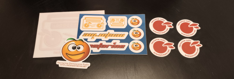Corner of a black table with one radio-design window cling, one sticker sheet, one large Necta sticker, and four fruit stickers with text around the edges arranged on top